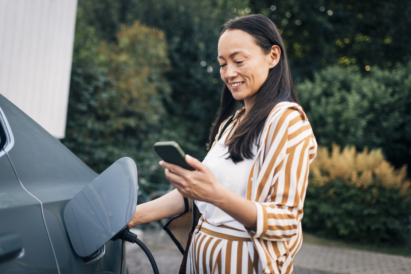 Fornøyd hovedbankkunde som lader elbil og bruker mobilbank på mobilen.