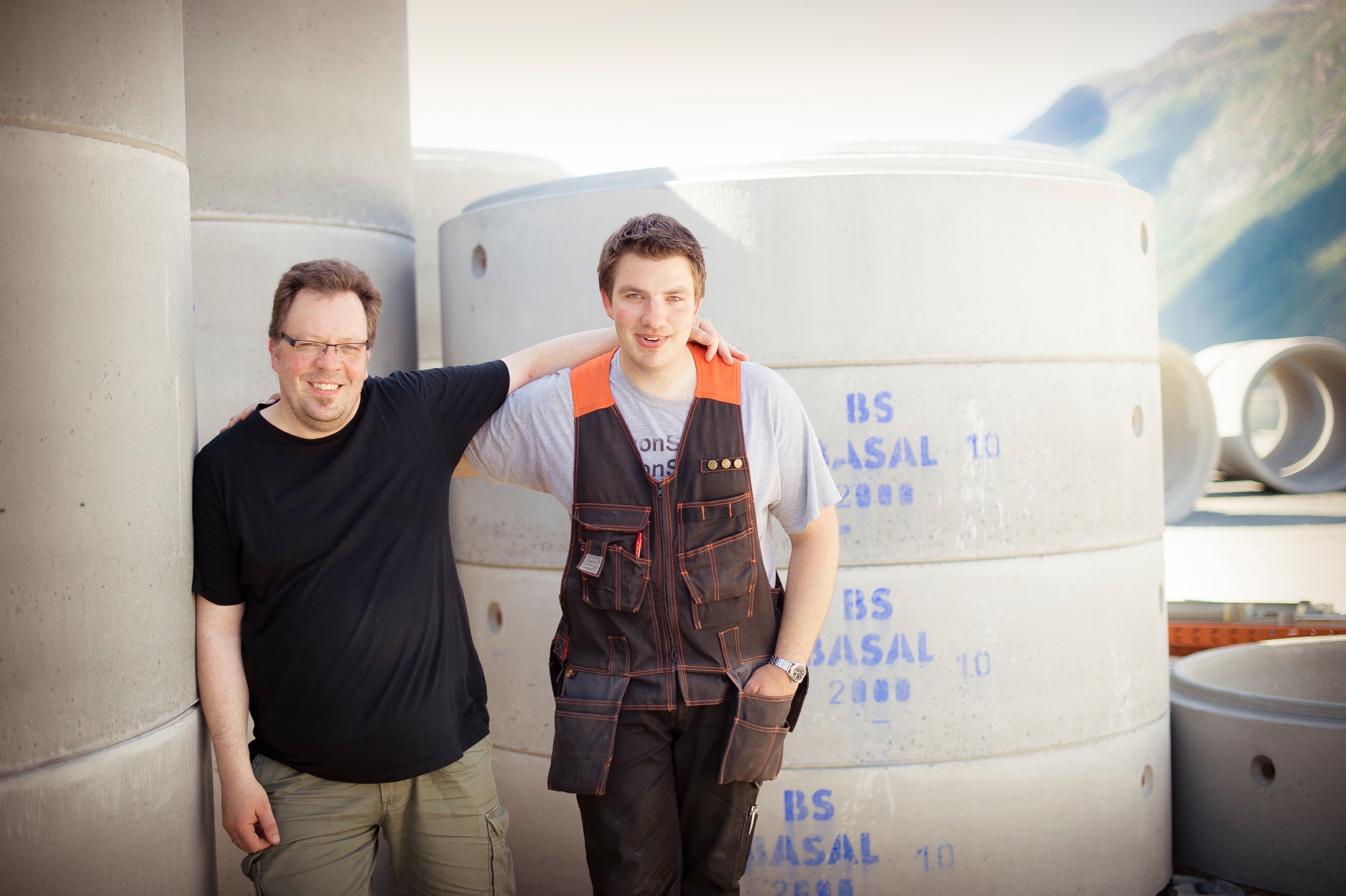 To arbeidskarer holder rundt hverandre foran store sylindre av betong