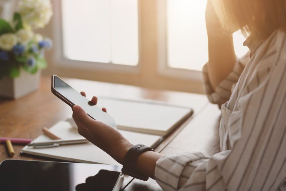 En dame holder en mobil i hånden. På bordet ligger et nettbrett og notisbok