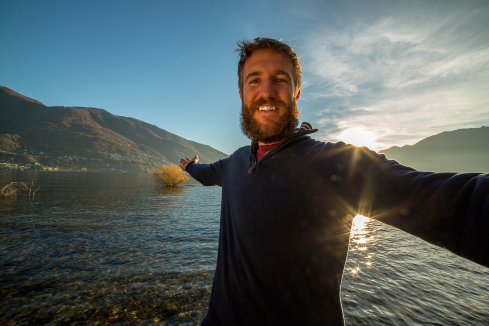 En mann strekker armene ut i havet. I bakgrunnen går solen ned i en fjord