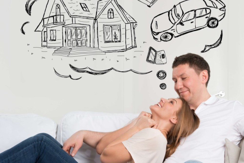 En kvinne ligger i fanget til en mann i en sofa. På veggen bak dem er det tegnet tenkebobler med bilde av et hus og en bil