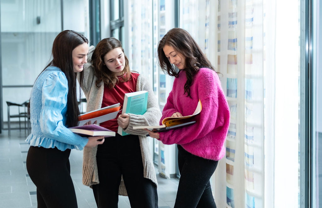 Tre kvinnelige studenter kikker på bøkene sine i gangen på Høgskulen