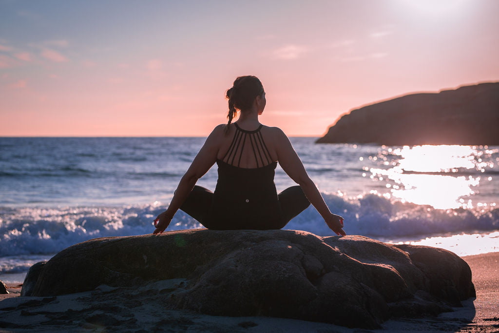 En dame sitter i yogaposisjon på en stein på stranden i soloppgang