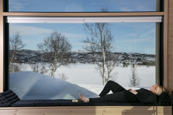 En kvinne ligger på en benk ved et stort vindu i en hytte. Ute er det et vakkert vinterlandskap.